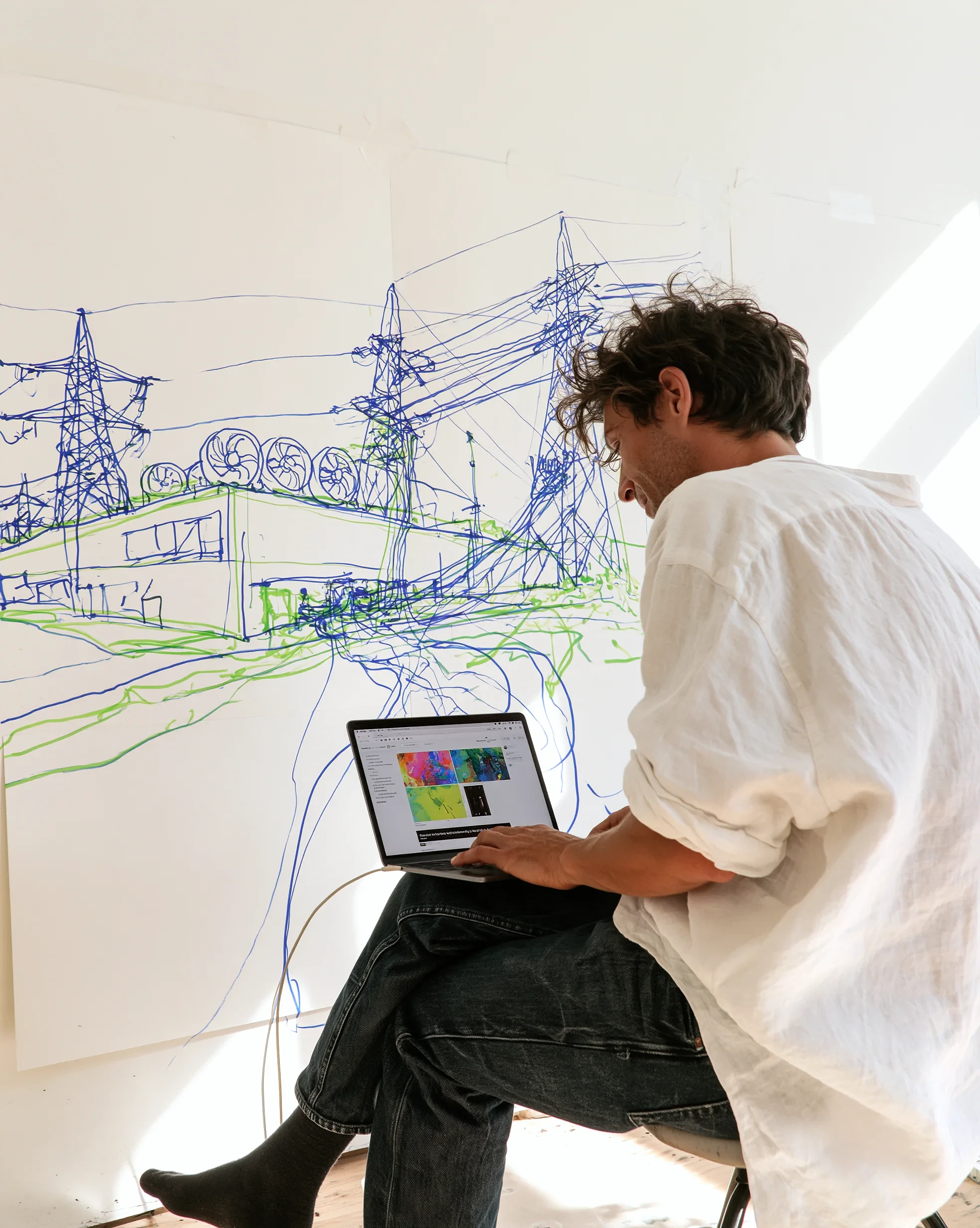 Electricity: a man in a white linen shirt sits on a stool with a laptop on his knees, in front of a wall drawing of pylons, power lines and a building with rows of fans.